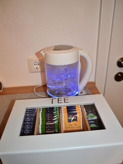Ein elektrischer Wasserkocher mit blauem Licht steht auf einem Tisch, während eine Teekiste mit verschiedenen Teesorten daneben liegt. Die Kiste ist mit "TEE" beschriftet.