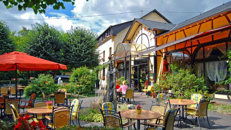 Ein einladendes Restaurant mit gemütlichen Sitzplätzen im Freien. Bunte Blumen und grüne Bäume umgeben die Terrasse.