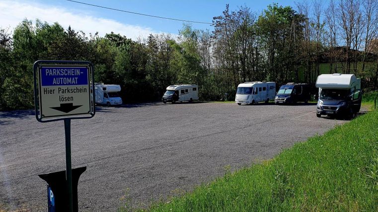 A parking lot for motorhomes with several vehicles. In the background, green trees and a clear sky can be seen.