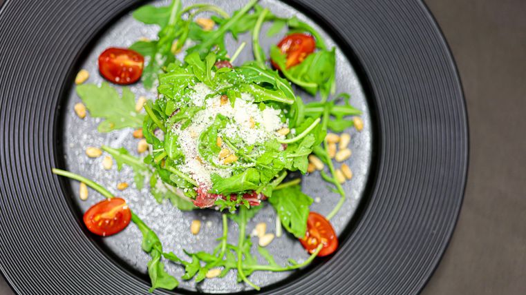 A fresh salad with arugula, cherry tomatoes, and pine nuts, served on a black plate. The salad is sprinkled with Parmesan and looks appetizing.