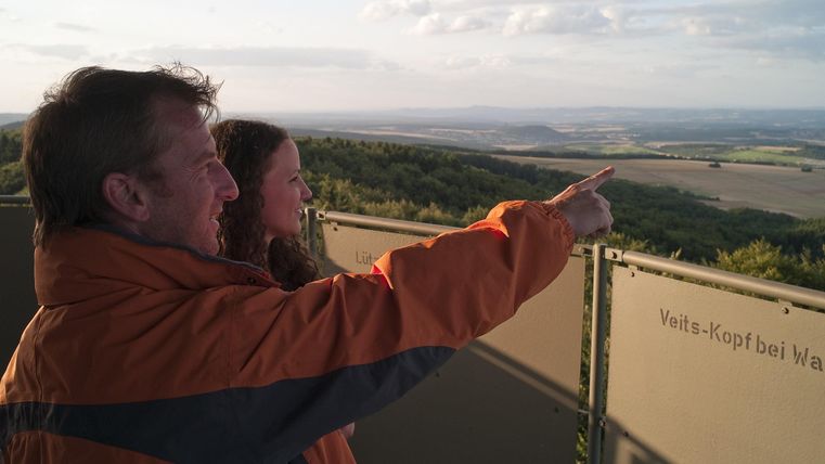 Ein Mann und eine Frau stehen an einem Aussichtspunkt und betrachten die Landschaft. Sie zeigen in die Ferne, während die Sonne untergeht.