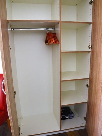 Ein leeres, moderner Kleiderschrank mit mehreren Fächern und einem Kleiderstangenbereich. Einige Holzkleiderbügel sind sichtbar, und unten befindet sich ein kleiner Safe.