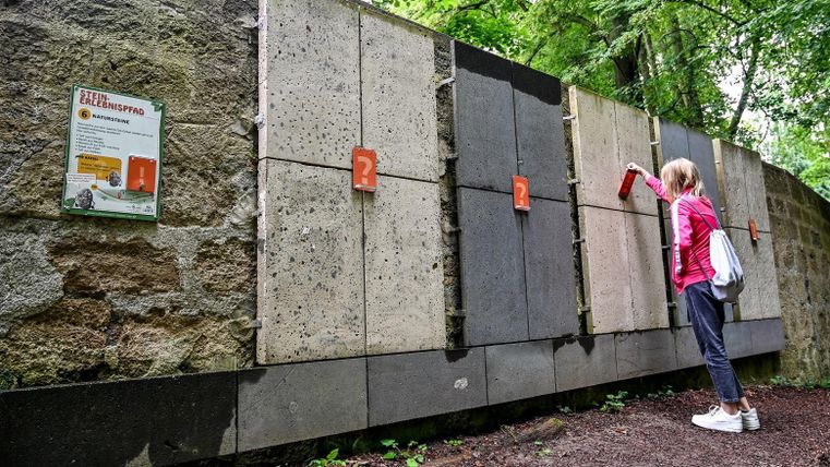 A person is standing in front of a wall with various concrete slabs. They are holding a red object in their hand and examining the slabs.