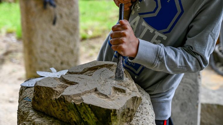 Eine Person schnitzt ein Muster in einen Steinblock. Das Motiv zeigt ein Blatt und wird mit einem Meißel bearbeitet.