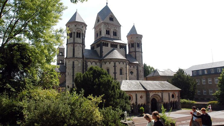 Eine historische Kirche mit mehreren Türmen, umgeben von Grünanlagen. Im Vordergrund sind einige Besucher zu sehen, die die Umgebung erkunden.