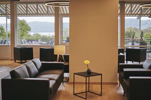 A modern lounge area with dark sofas and a small table. Large windows provide a view of the nature outside.
