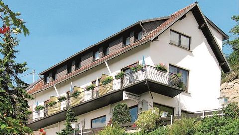 Ein malerisches Haus mit mehreren Fenstern und einem Balkon. Es ist von grünen Pflanzen umgeben und hat einen klaren blauen Himmel im Hintergrund.