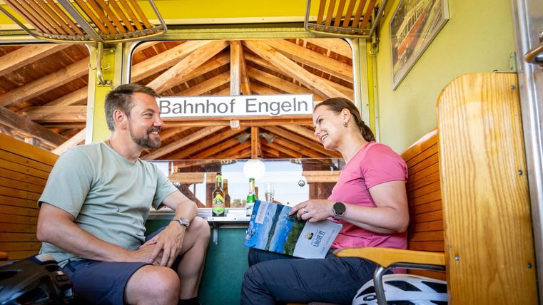 A man and a woman are sitting in a carriage and talking to each other. In the background, the sign "Engeln Station" is visible.
