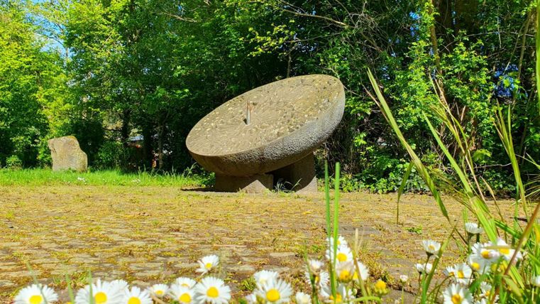 Ein runder, steinerner Obelisk steht in einer grünen Umgebung. Im Vordergrund blühen Gänseblümchen.