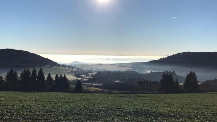 Eine weite Landschaft unter einem klaren Himmel mit der Sonne im Hintergrund. Nebel liegt über den Hügeln und Wäldern.