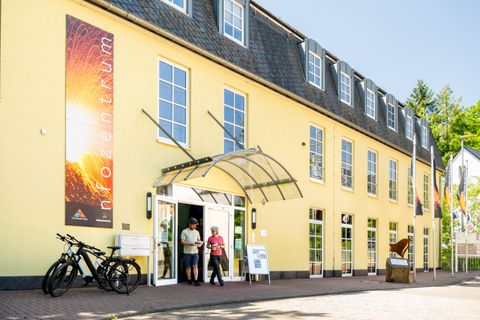 Ein modernes Informationszentrum mit gelber Fassade und großen Fenstern. Zwei Personen treten durch den Eingang, während Fahrräder davor stehen.