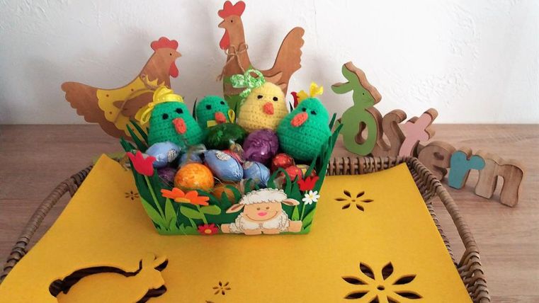Ein buntes Osternest mit gehäkelten Küken und Schokoladen-Eiern. Im Hintergrund steht eine Holzdekoration mit dem Schriftzug "Ostern".