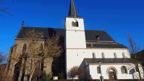 An impressive church with a tall tower against a clear blue sky. The architecture combines different styles and is surrounded by trees.