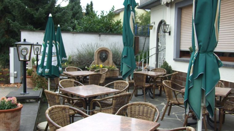 Ein gemütlicher Außenbereich mit Rattanstühlen und Tischen. Grüne Sonnenschirme spenden Schatten und es gibt eine kleine Gartenlandschaft.