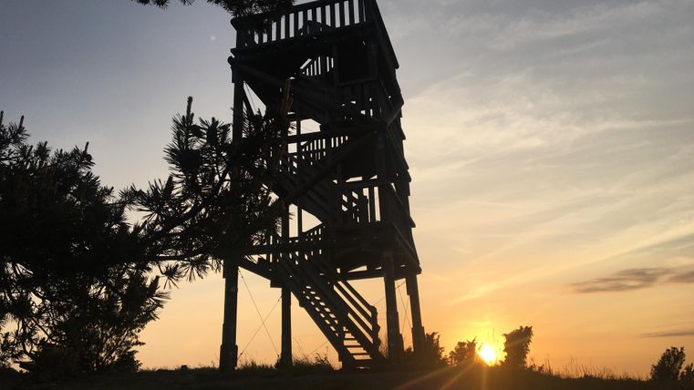 Een uitkijktoren staat silhouet tegen de zonsondergang. De lucht is oranje en roze gekleurd, terwijl het licht mild over het landschap straalt.
