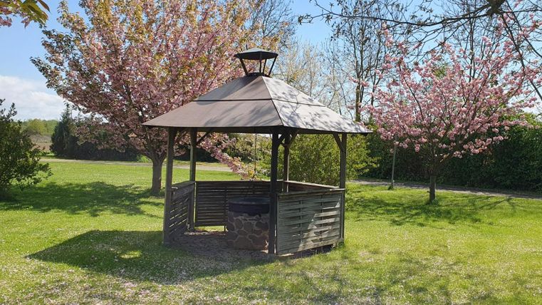 Ein gemütlicher Pavillon steht in einer grünen Wiese. Im Hintergrund blühen rosa Bäume und der Himmel ist blau.