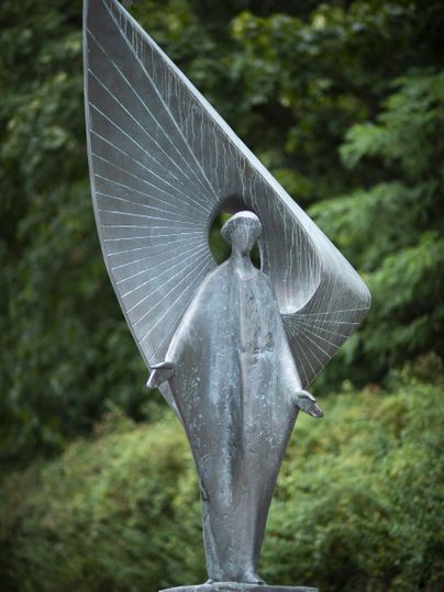 A statue of an angel with large, curved wings stands in nature. The figure has a calm aura and is surrounded by green trees.