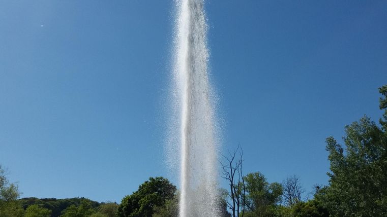 Ein Wasserfontäne sprudelt hoch in den blauen Himmel. Umgeben von grünen Bäumen und roten Steinen ist die Szenerie beruhigend.