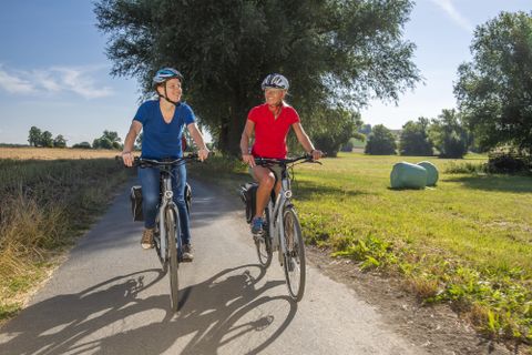 Zwei Radfahrerinnen fahren lächelnd einen Radweg entlang. Im Hintergrund sind grüne Wiesen und Bäume sichtbar.