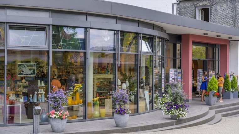 Ein modernes Geschäft mit großen Fenstern und einer einladenden Außenanlage. Menschen betreten und verlassen den Laden, umgeben von bunten Blumen.