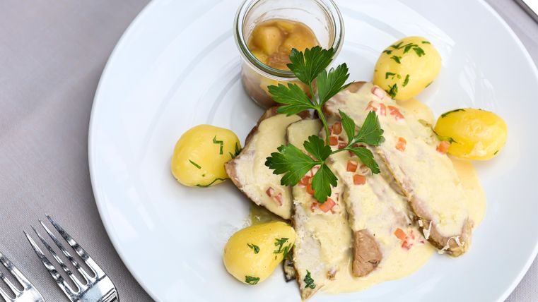 Ein schön angerichteter Teller mit zartem Fleisch in einer cremigen Sauce, umgeben von Kartoffeln und einem Glas mit einer süßen Beilage. Frische Petersilie verleiht dem Gericht einen eleganten Akzent.