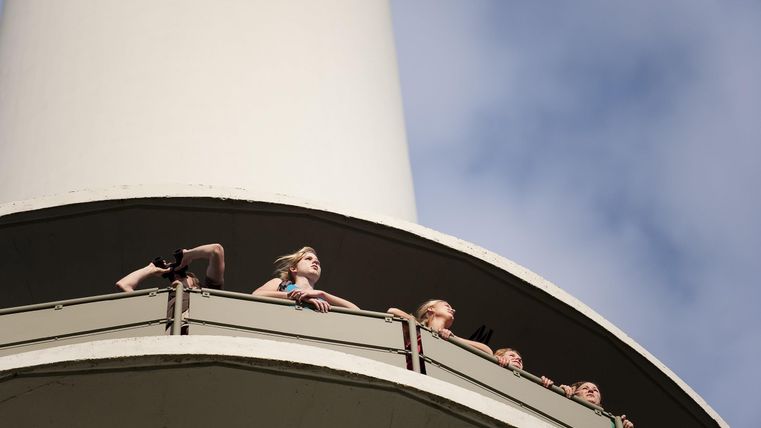 Eine Gruppe von Menschen steht auf einem Balkon und schaut nach oben. Der Himmel ist blau mit einigen Wolken.