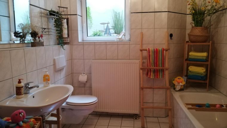 A modern bathroom with white tiles and large windows. There is a bathtub, a sink, and colorful towels on a shelf.