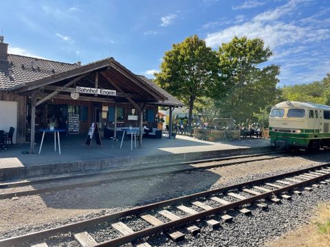A train station with a wooden building and a train on the tracks. The surroundings are green with trees and a blue sky.