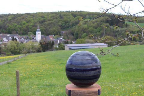 Eine idyllische Landschaft mit einem großen See und umgebenden Wäldern. Im Vordergrund sind Felder und ein Gebäude sichtbar.
