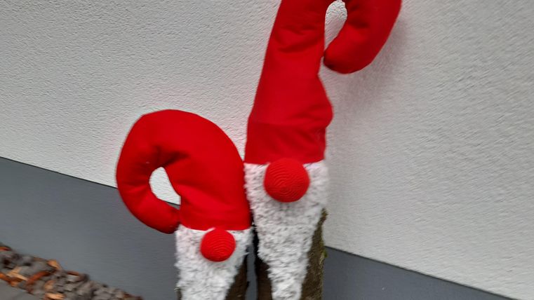 Zwei dekorierte Baumstämme mit roten Weihnachtsmützen und einem weißen Bart. Sie sind mit einer roten Schleife verziert und stehen vor einer weißen Wand.