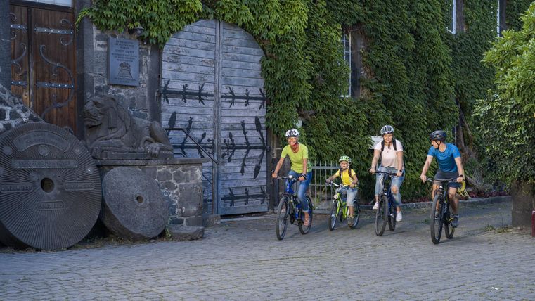 Eine Familie fährt mit Fahrrädern auf einem gepflasterten Weg. Im Hintergrund sieht man eine grüne Wand aus Pflanzen und große, alte Steine.