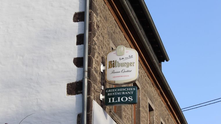 Ein Schild eines griechischen Restaurants namens "Ilios". Das Restaurant befindet sich an einer älteren, gemauerten Wand.