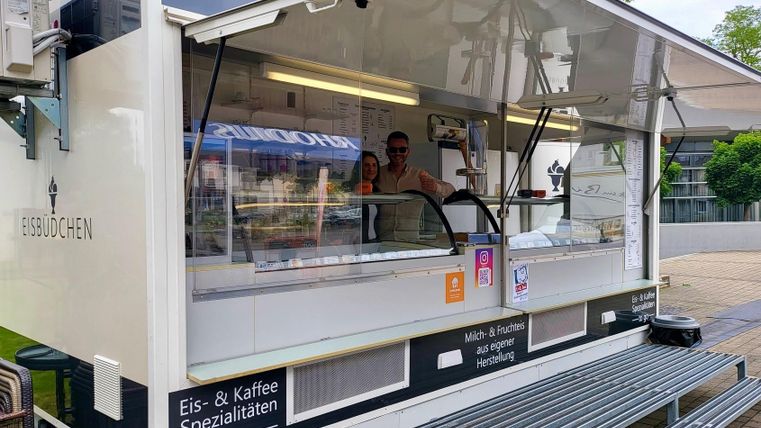 An ice cream stand with a modern, white sales trailer. The sellers stand behind the counter and offer ice cream and coffee specialties.