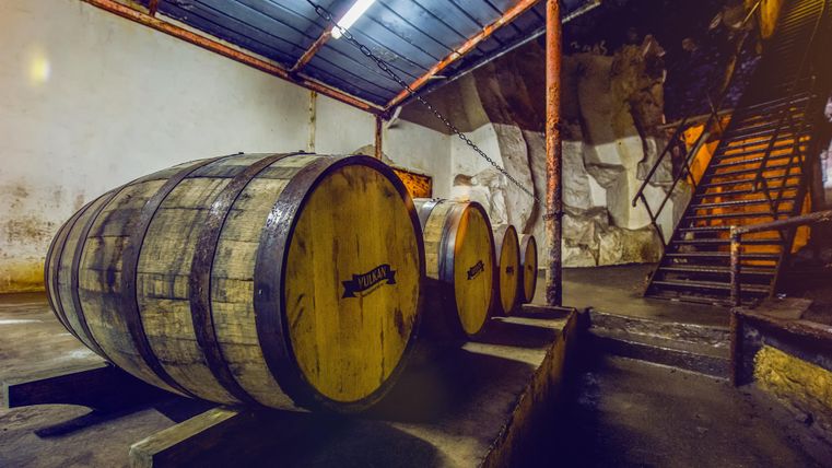 Ein Innenraum mit großen Holzfässern, die zum Lagern von Wein genutzt werden. Im Hintergrund ist eine Treppe sichtbar, die nach oben führt.