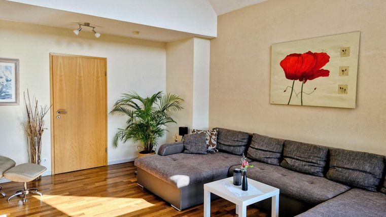 Ein modernes Wohnzimmer mit einem großen grauen Sofa und einem weißen Tisch. An der Wand hängt ein Bild mit einer roten Blume, und es gibt eine Pflanze in der Ecke.