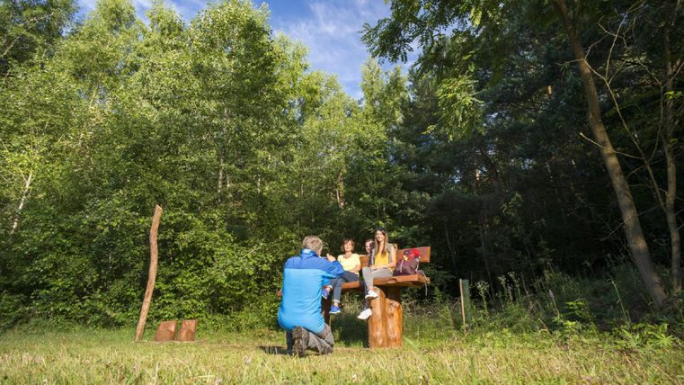 Eine Gruppe von drei Personen sitzt auf einer Bank im Wald. Ein Fotograf kniet davor und macht ein Foto.