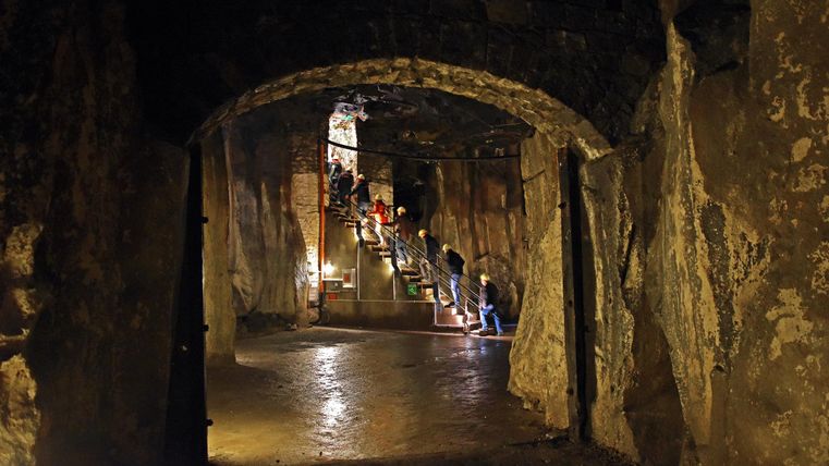 Ein unterirdischer Raum mit Steinwänden und einer Treppe im Hintergrund. Das Licht beleuchtet den Weg und zeigt Menschen, die hinuntergehen.