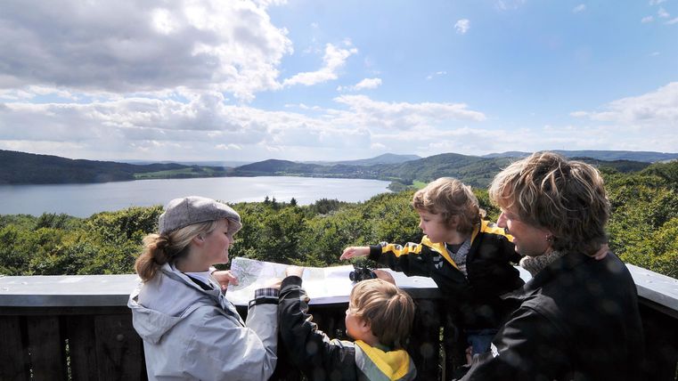 Eine Familie steht an einem Aussichtspunkt und schaut auf eine schöne Landschaft. Der Himmel ist teilweise bewölkt und der See ist im Hintergrund zu sehen.