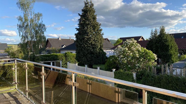 Ein schöner Ausblick von einem Balkon auf grüne Gärten und Häuser. Der Himmel ist blau mit einigen Wolken.