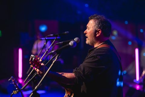 Ein Musiker spielt Gitarre auf einer Bühne. Bunte Lichter sorgen für eine stimmungsvolle Atmosphäre.