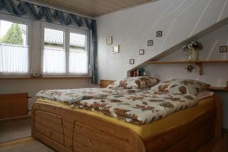 Ein gemütliches Schlafzimmer mit einem Holzbett und bunten Bettdecken. Große Fenster lassen viel Tageslicht herein.