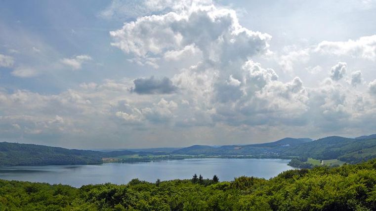 Ein malerischer See umgeben von grünen Wäldern und sanften Hügeln. Der Himmel ist teils bewölkt und strahlt eine friedliche Atmosphäre aus.