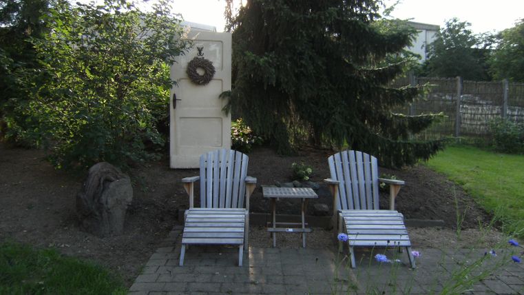 Ein gemütlicher Garten mit zwei Liegestühlen und einem kleinen Tisch. Im Hintergrund steht eine Tür, umgeben von Bäumen und Pflanzen.