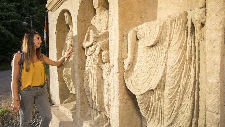 Eine Frau berührt historische Skulpturen an einer Wand. Die Figuren zeigen verschiedene kunstvolle Darstellungen.