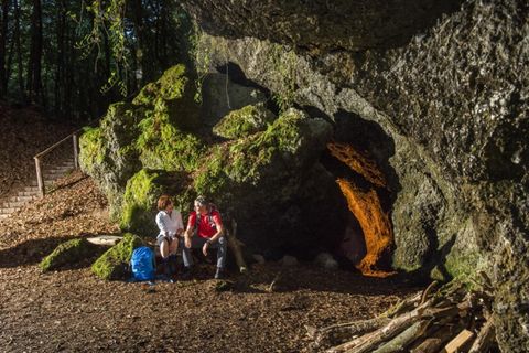 Eine Gruppe von Menschen sitzt entspannt vor einer großen, moosbedeckten Felsformation. Im Hintergrund ist eine Höhle mit orangefarbenem Licht sichtbar.