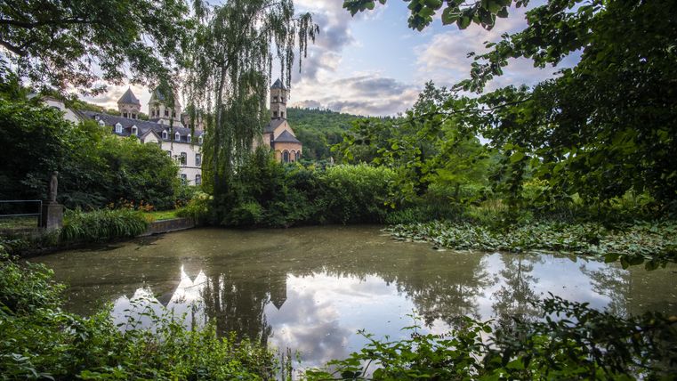 A serene pond surrounded by lush greenery and trees. In the background, a historic building can be seen, reflecting in the water.