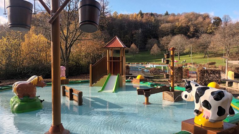 Ein Wasserspielplatz mit einer Rutsche und lustigen Tieren. Die Umgebung ist grün und die Sonne scheint.