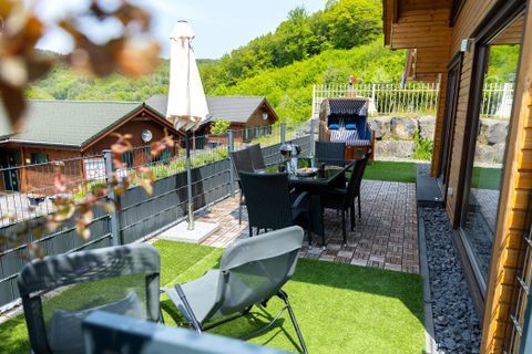 Eine gemütliche Terrasse mit Stühlen und einem Tisch, umgeben von grüner Landschaft. Im Hintergrund sind Ferienhütten sichtbar.