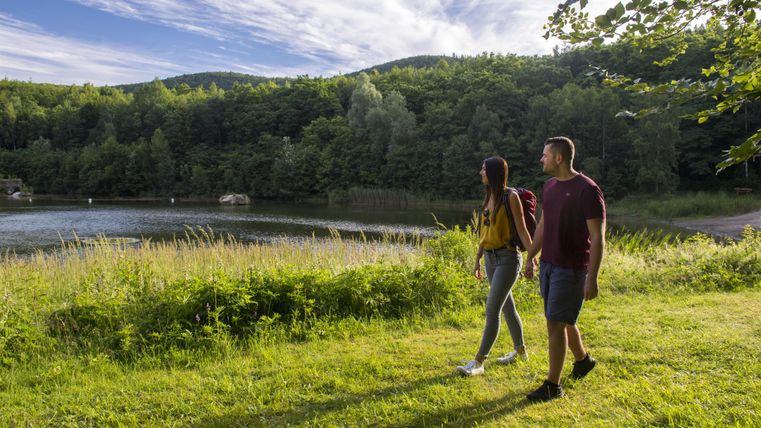 A couple walks along the shore of a calm body of water. Surrounded by green mountains and a clear sky, they enjoy nature.