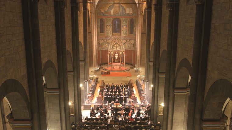 Eine beeindruckende Innenansicht einer Kirche mit hohen Decken und schönem Licht. Im Vordergrund ist ein Orchester zu sehen, das vor einem festlichen Altar spielt.